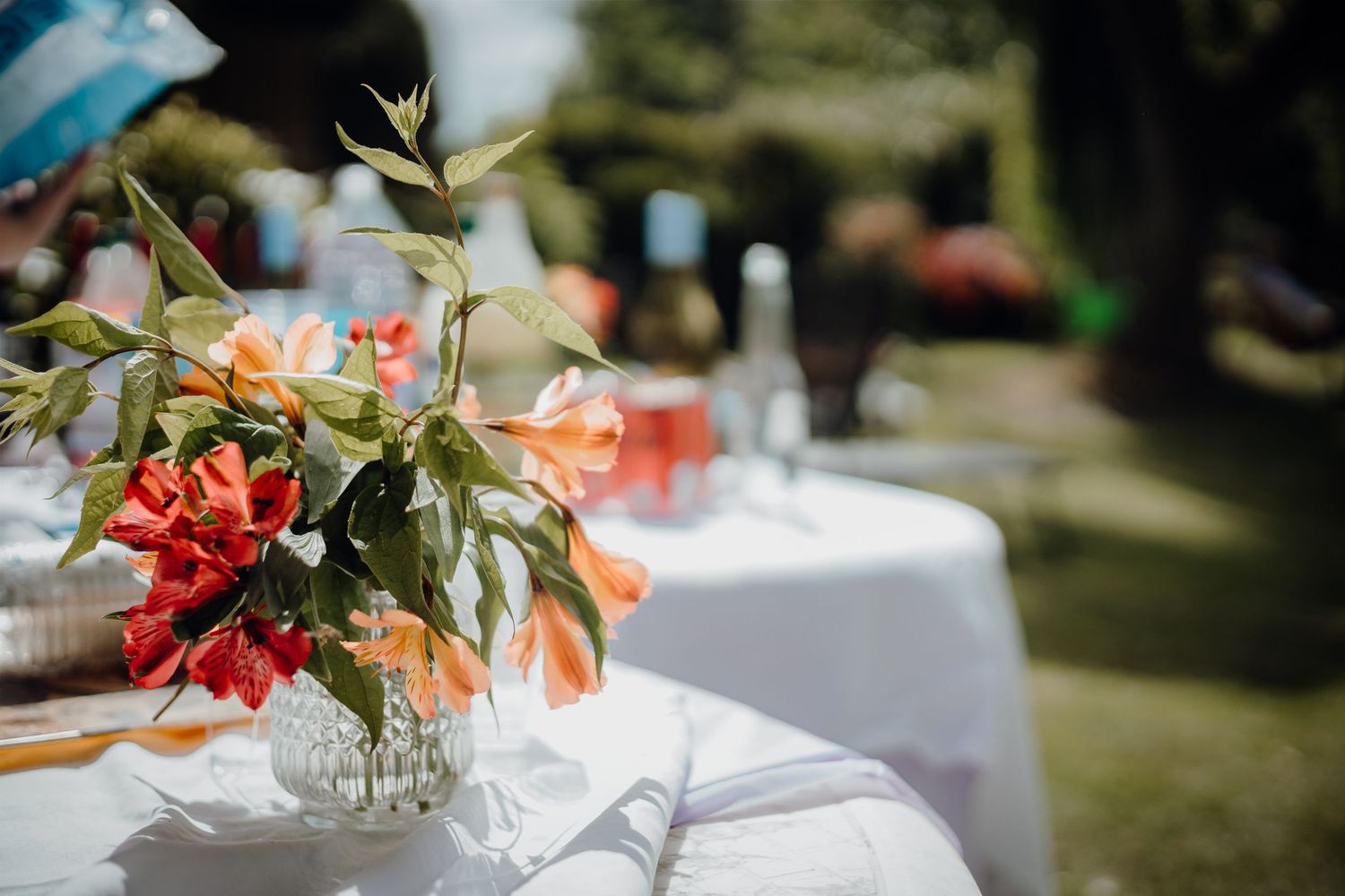 Elegant floral centerpieces with orange and red blooms on white tablecloths at outdoor event.