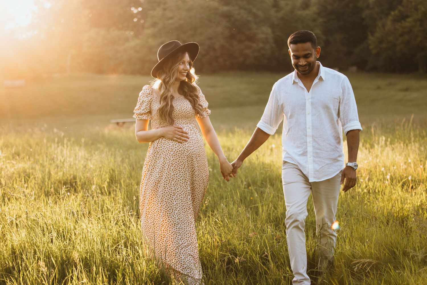 A couple walks together through a golden sunlit meadow during sunset wearing casual white and beige outfits.