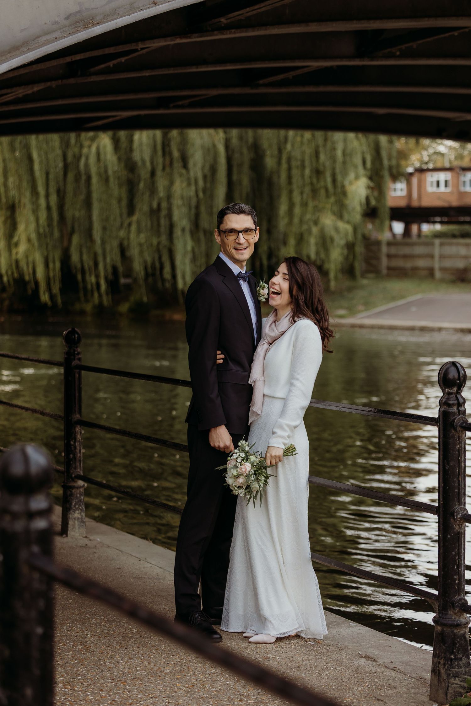 Couple sharing tender moment by riverside with weeping willow trees in background.