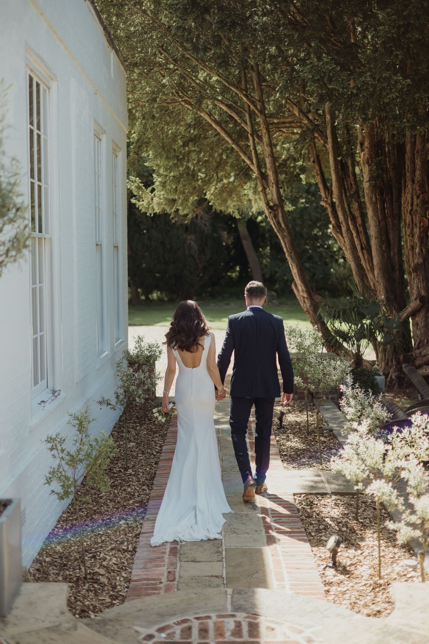 Newlyweds walking along a white building pathway surrounded by lush greenery.