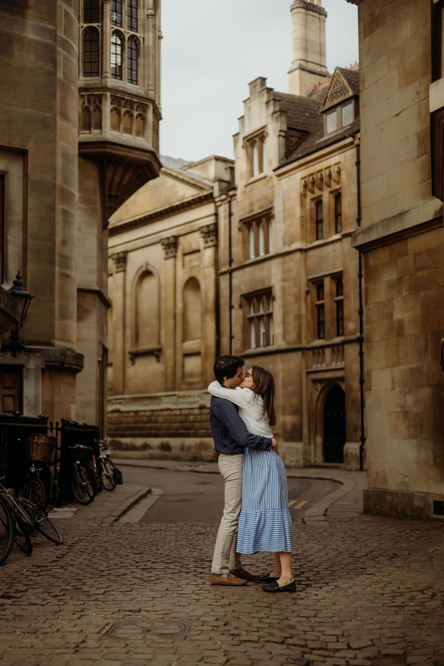A couple embraces on a cobblestone street surrounded by historic university buildings.