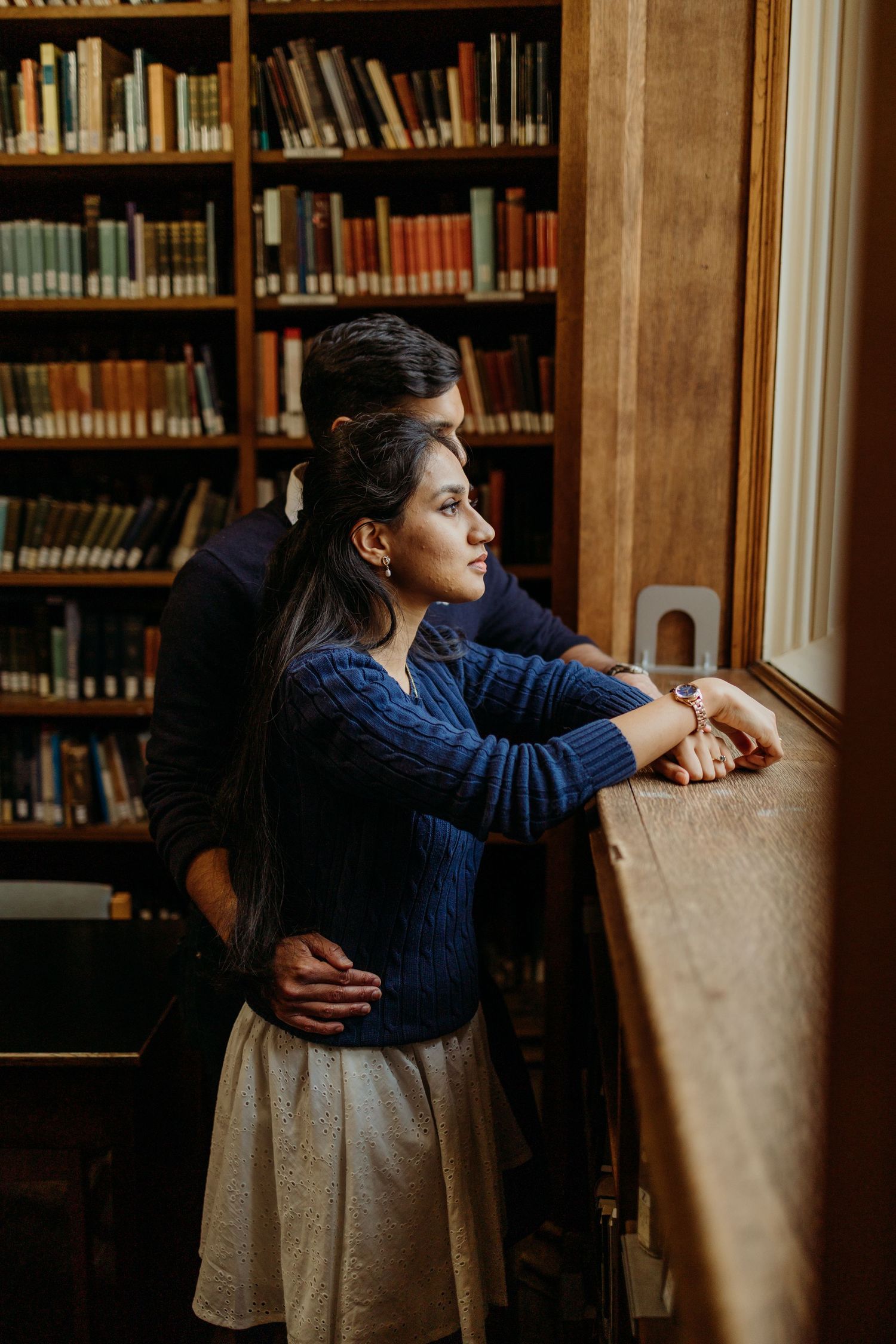 A person in navy blue sits contemplatively by a window in a library setting.