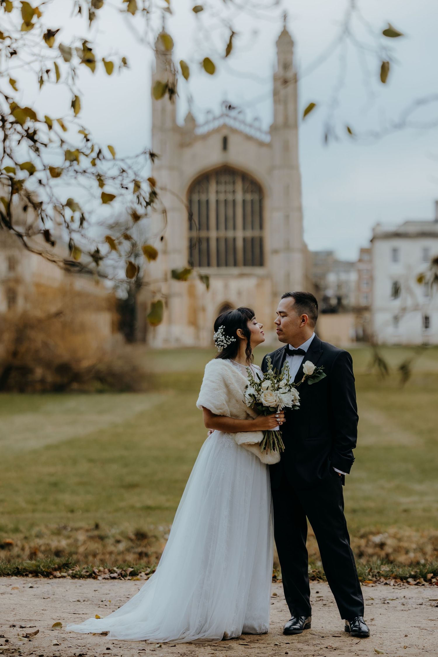 A wedding couple poses in front of a grand gothic architecture building with autumn leaves.