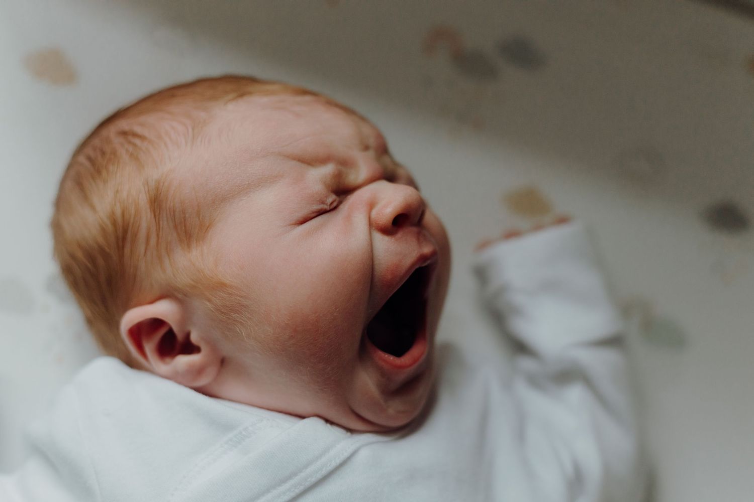 Newborn baby yawning while lying on white bedding in natural light.