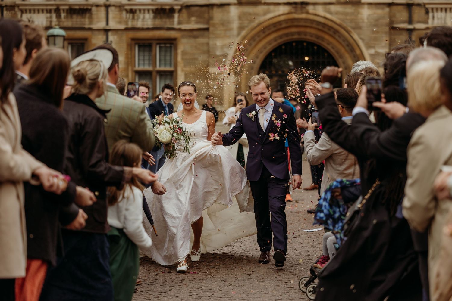 Guests toss flower petals at a wedding ceremony outside a historic stone building.