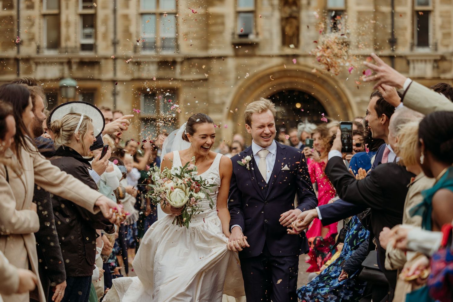 Joyful wedding celebration with guests throwing confetti at a grand stone building entrance.
