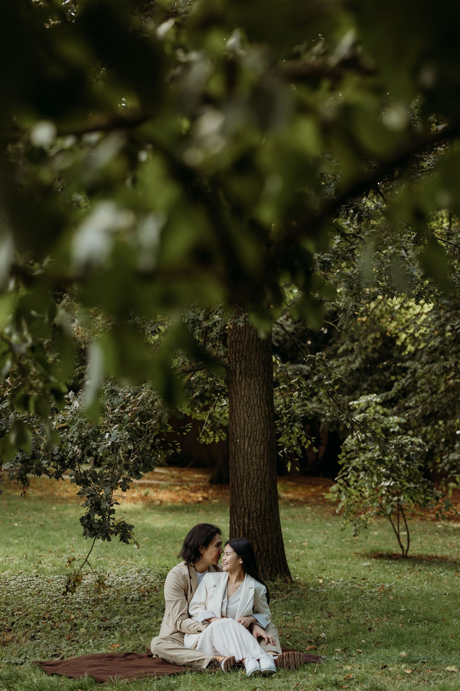 A couple sits beneath large trees in a lush garden setting.
