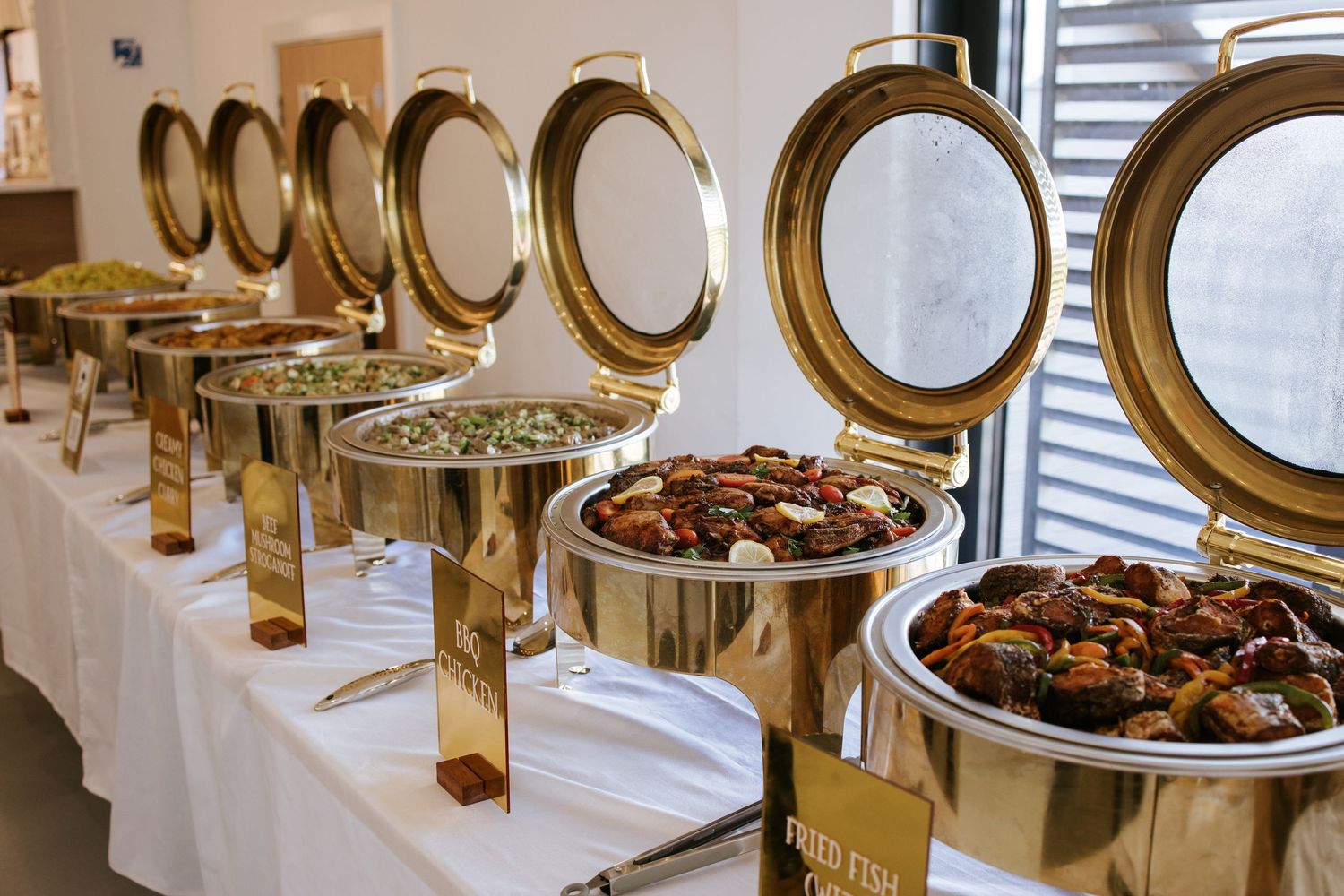Buffet setup with shiny gold chafing dishes and white tablecloths at an elegant event.