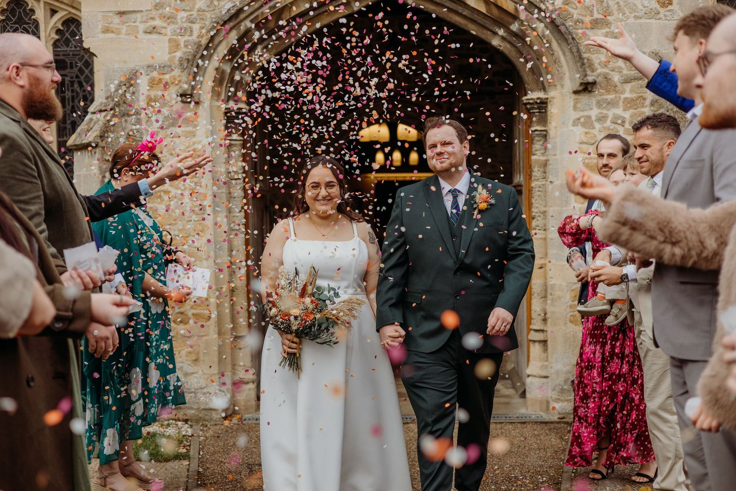 Wedding guests throw confetti outside a historic stone church during a celebration.