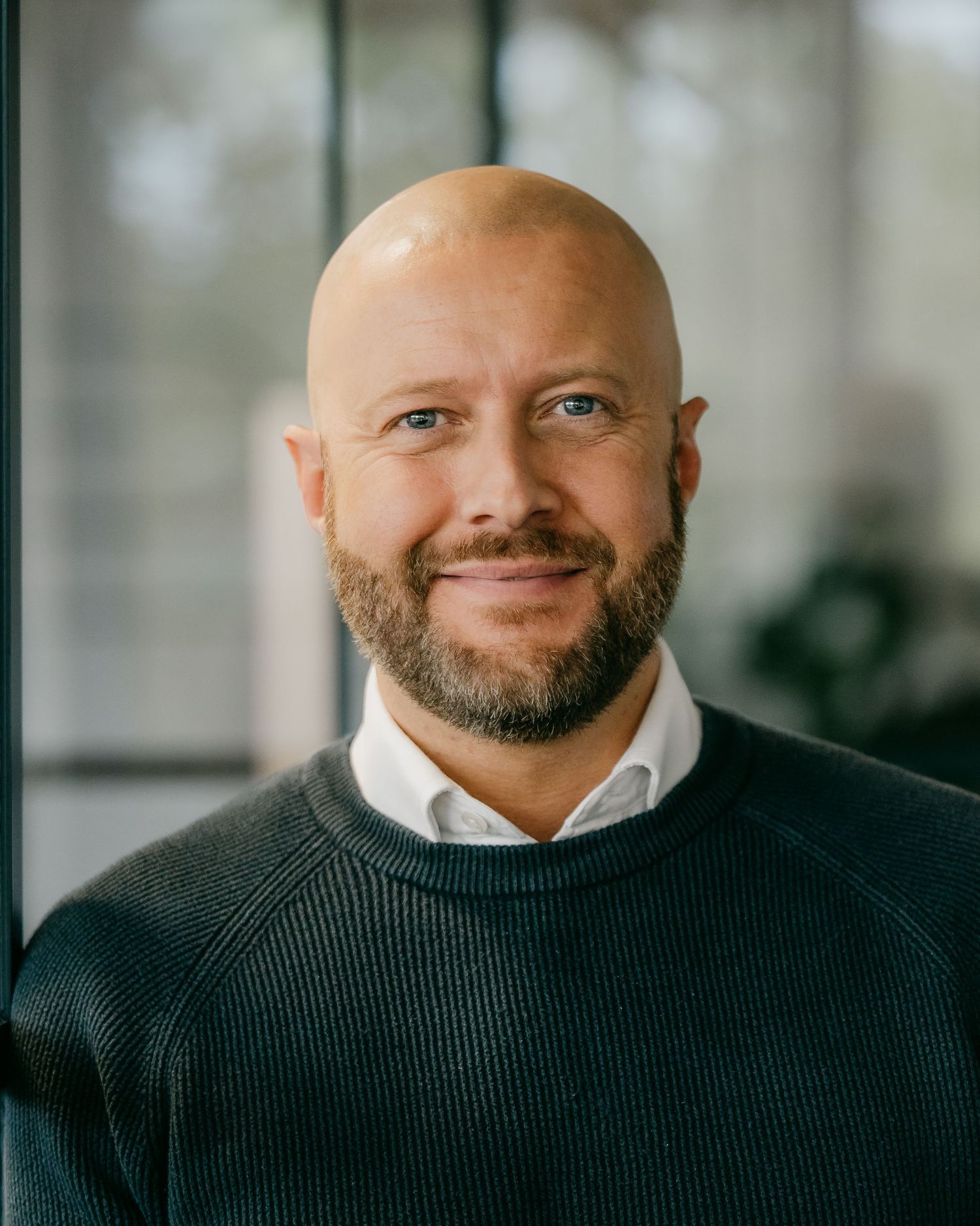 Professional headshot of someone in a black sweater smiling warmly against a bright office background.