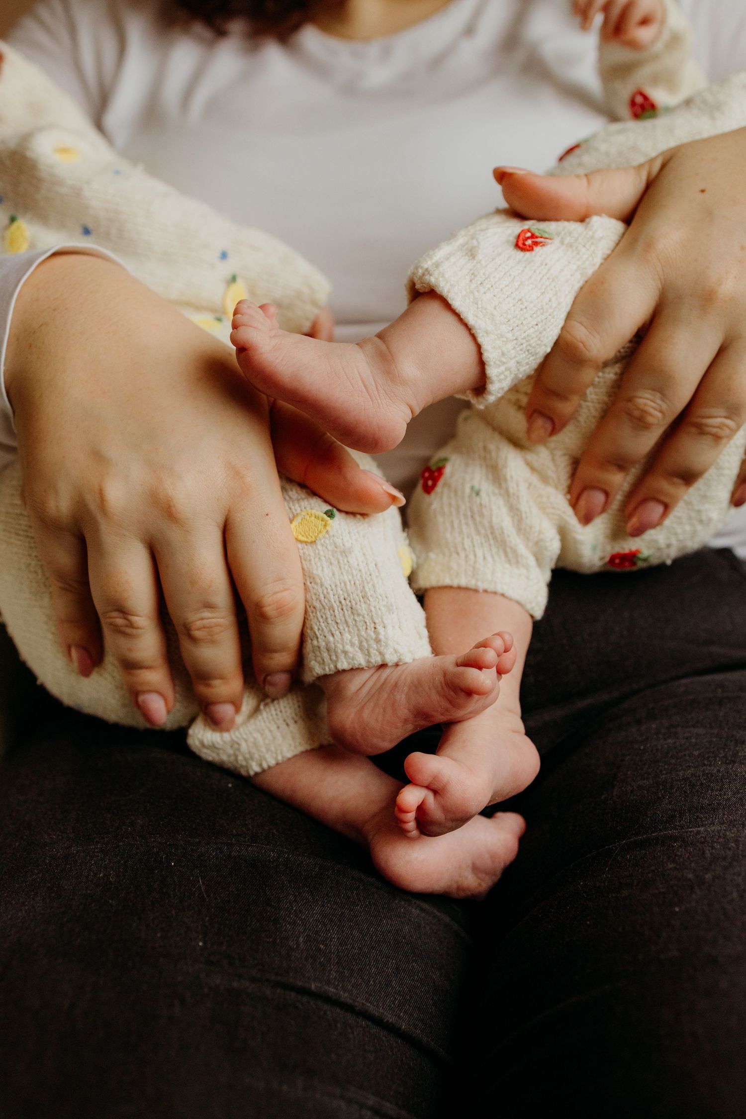 Close-up series of tiny baby feet being held by gentle hands wearing a cream knit sweater against dark fabric.