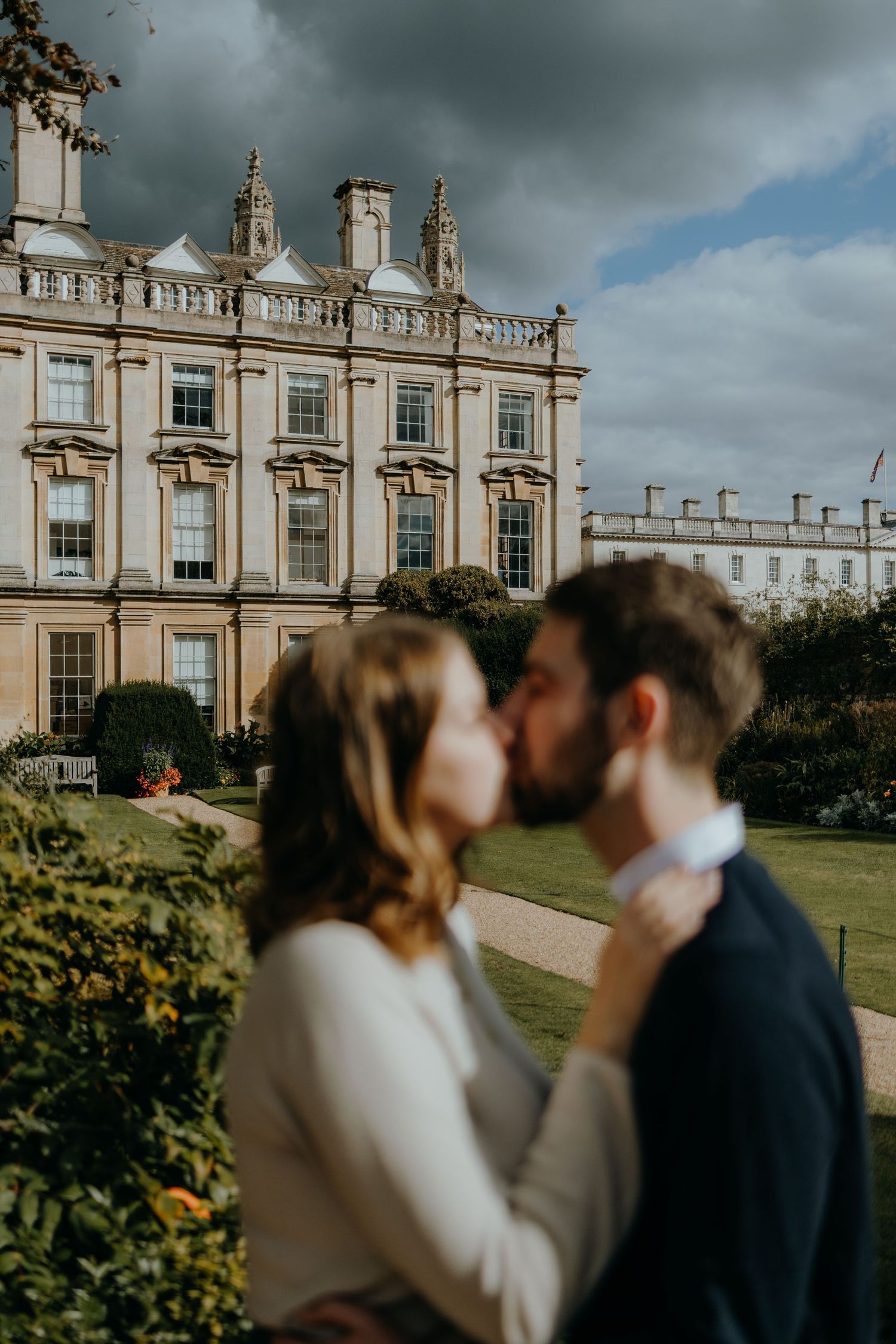 Wedding couple embracing in front of a grand historic mansion with dramatic cloudy sky.