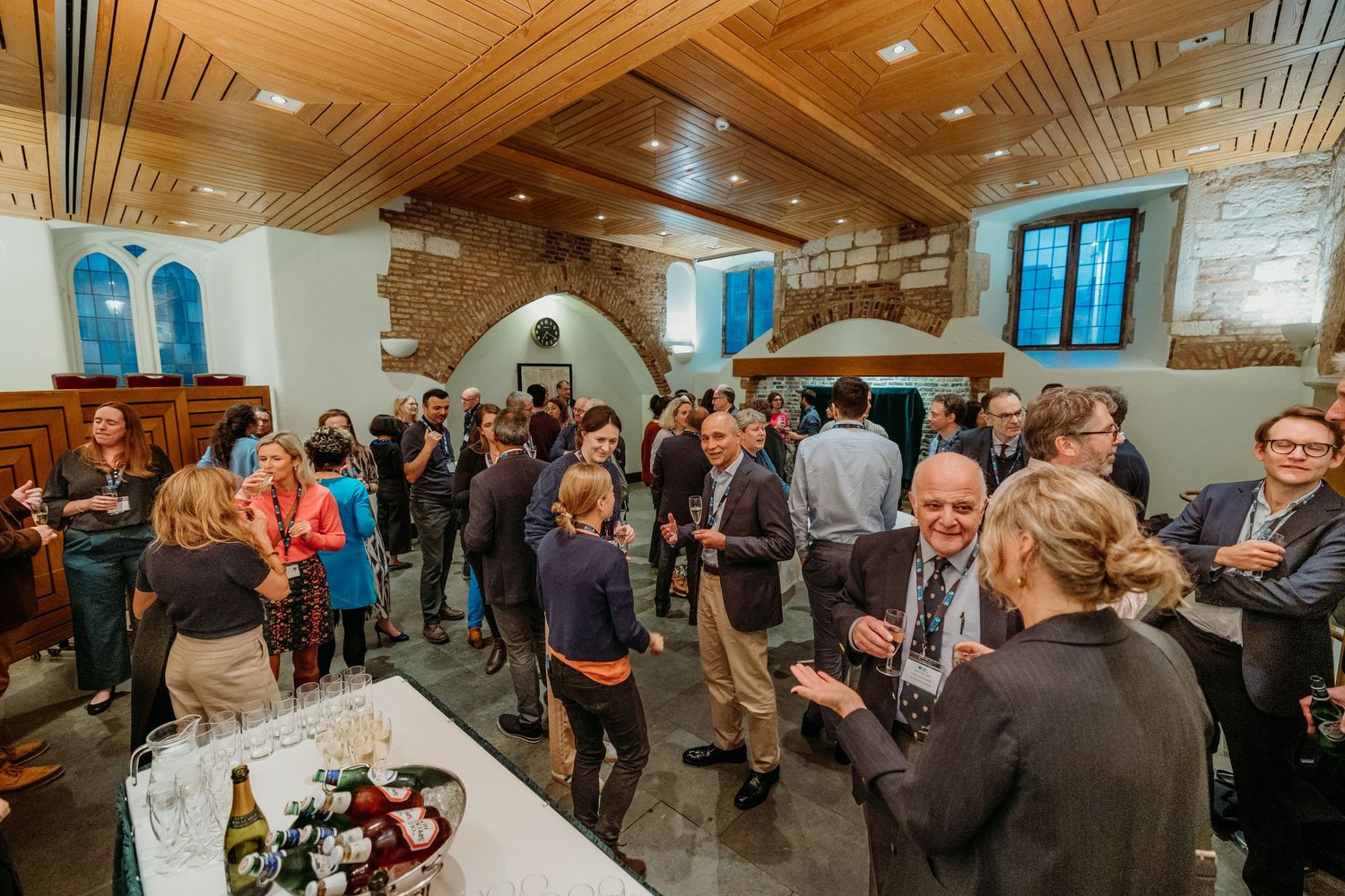 Large group mingling at an indoor reception with wooden ceiling and bright lighting.