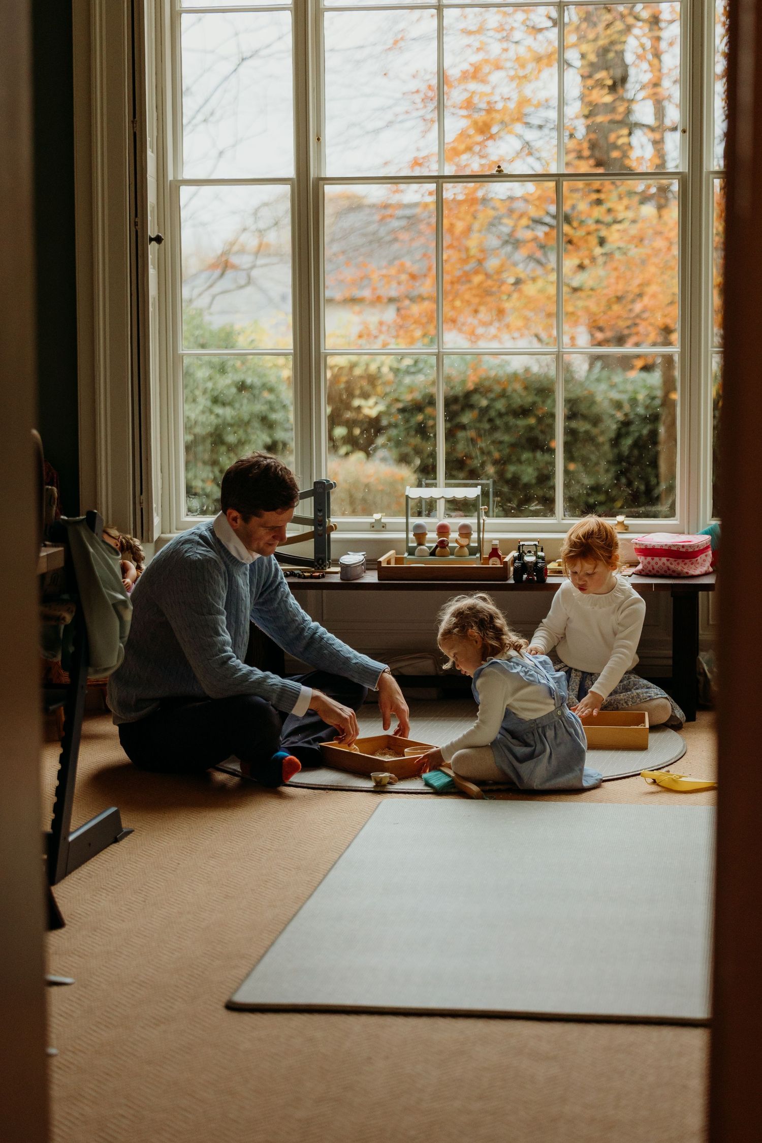 Family reading together by large windows with autumn foliage visible outside.