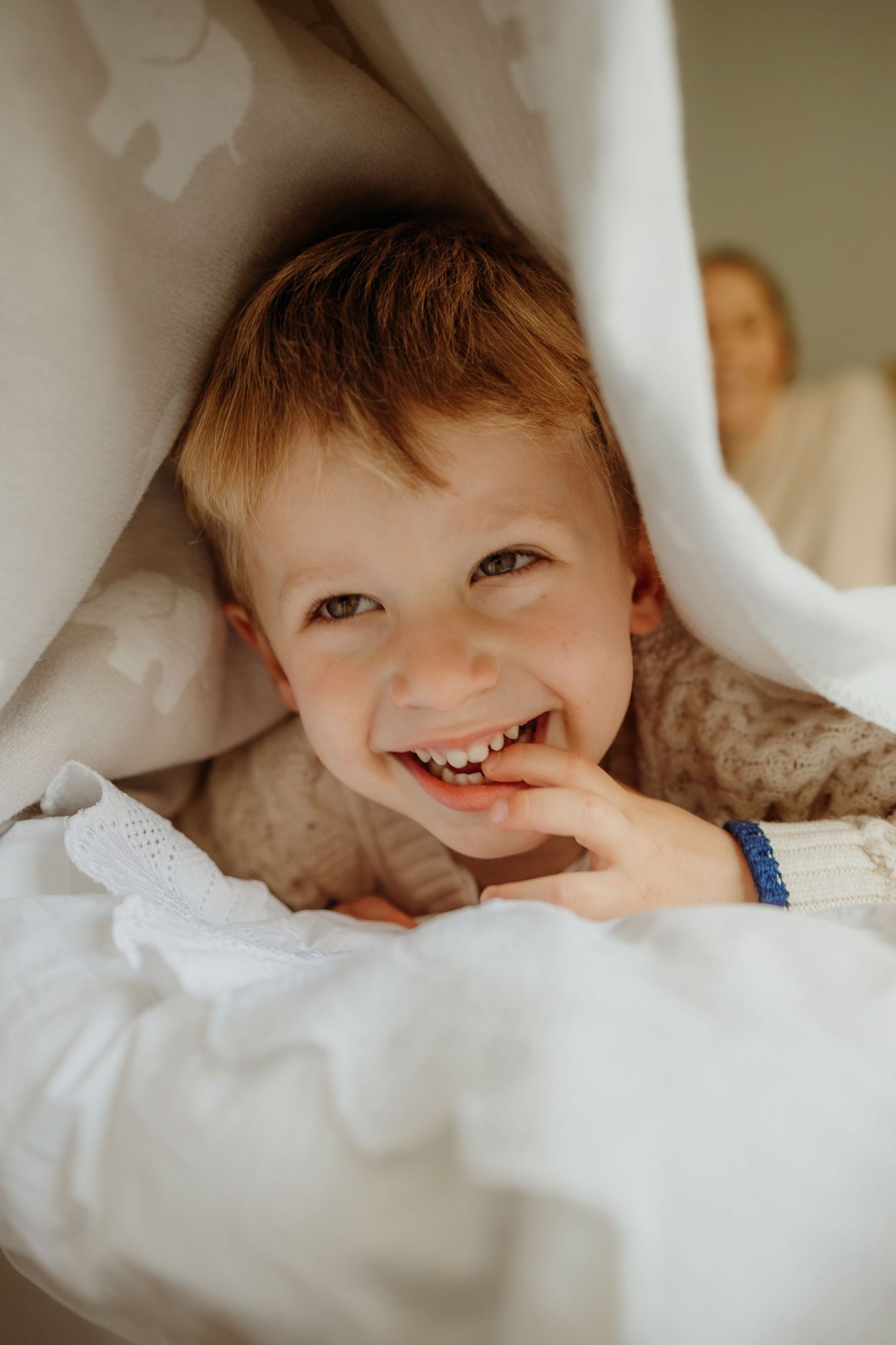 Child peeking out from under white blankets with a joyful smile.