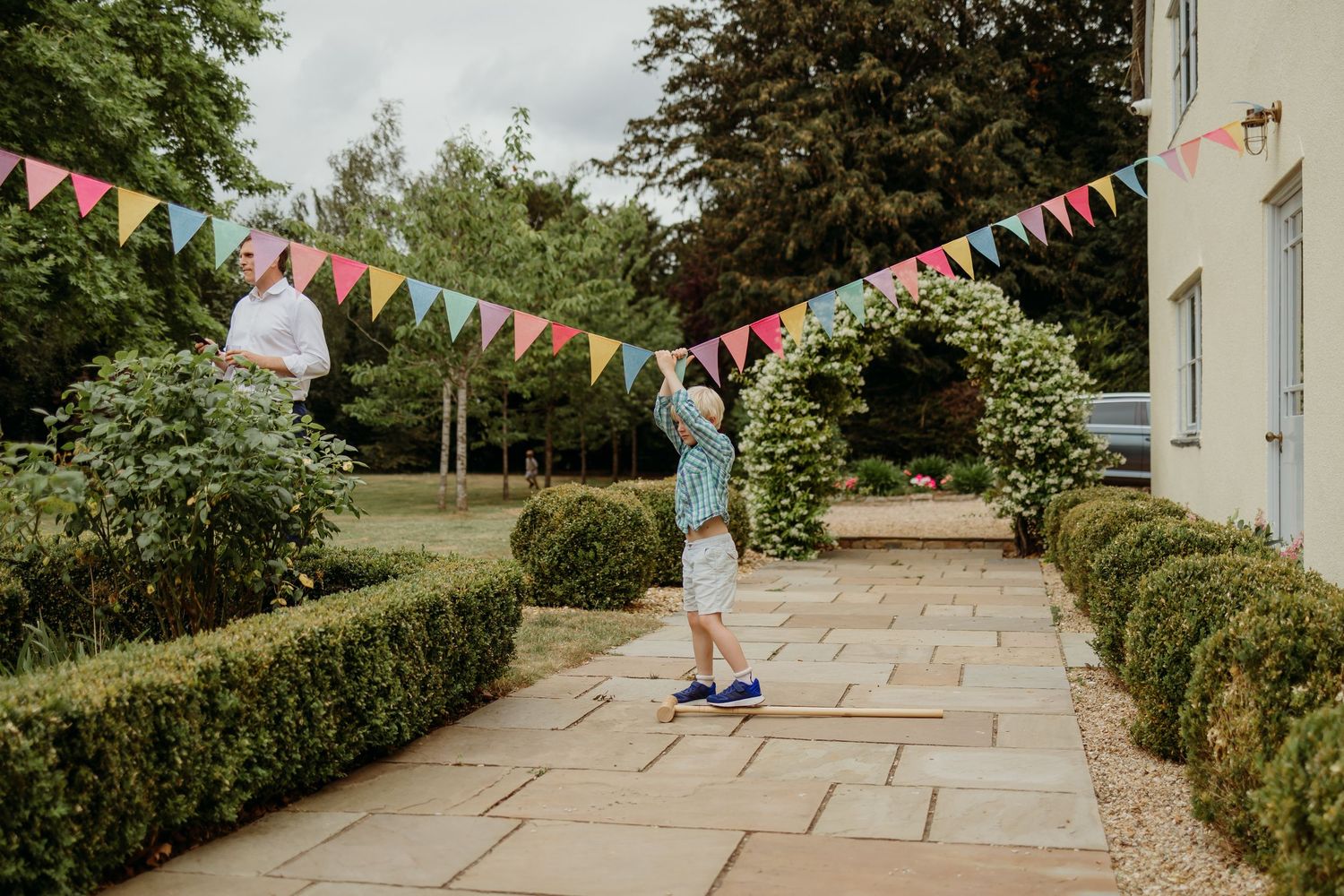 Colorful triangle bunting decorates an outdoor pathway with hedges and buildings.