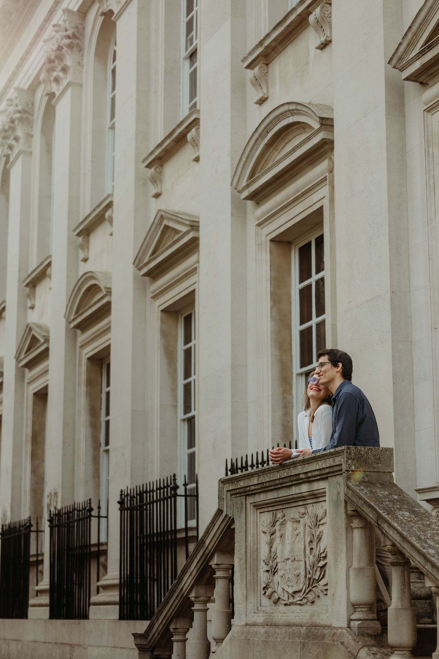 A romantic couple shares an intimate moment on a grand balcony with classical architecture in the background.