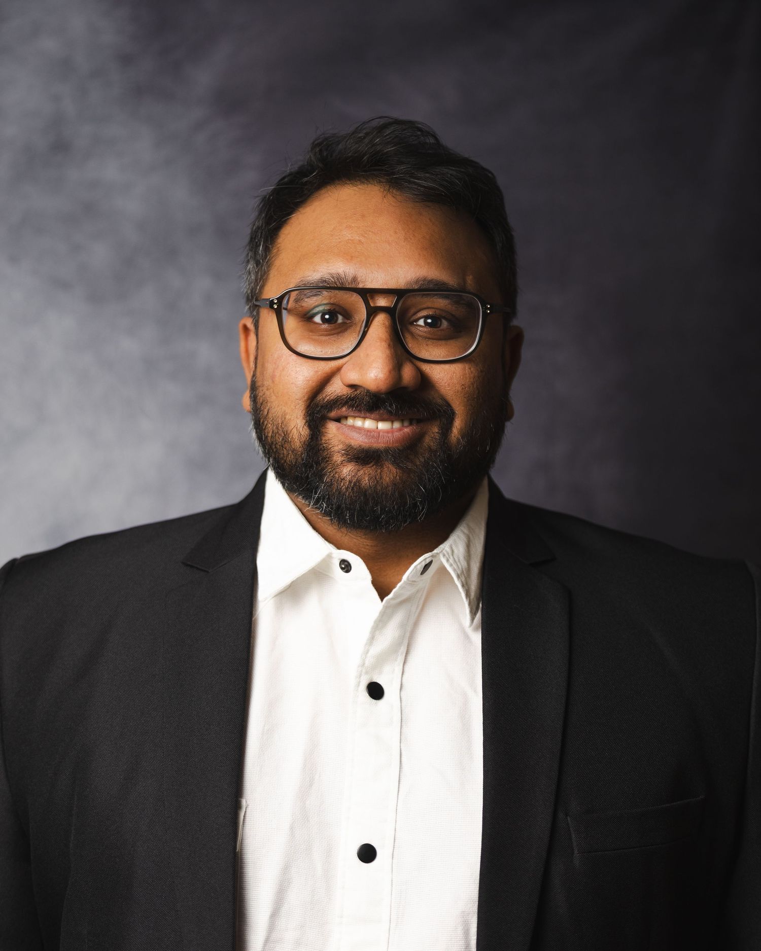 Professional headshot of businessman in suit and glasses against dark background.