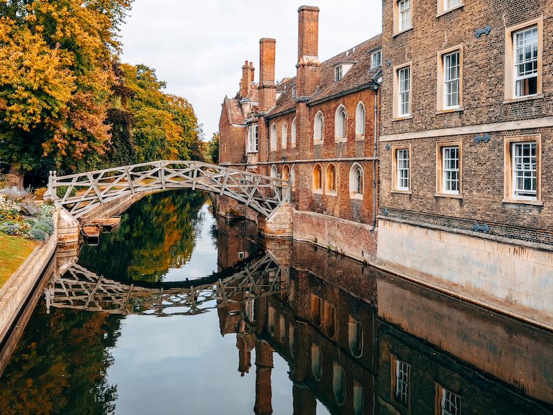 Historic brick buildings line a scenic canal in Cambridge with autumn trees reflecting golden colors in the water.