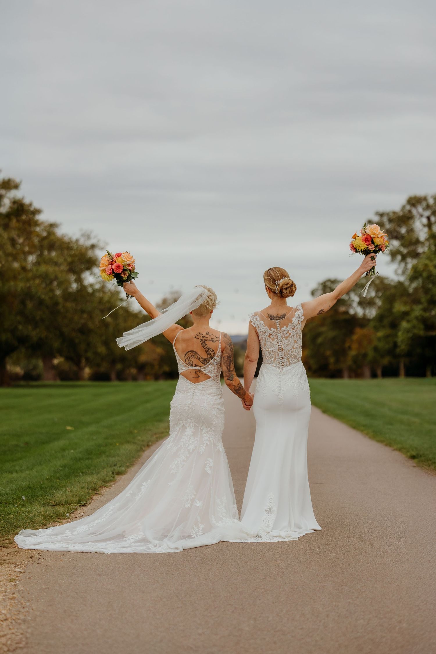 Two brides in lace wedding dresses raising bouquets on a path surrounded by green lawn.