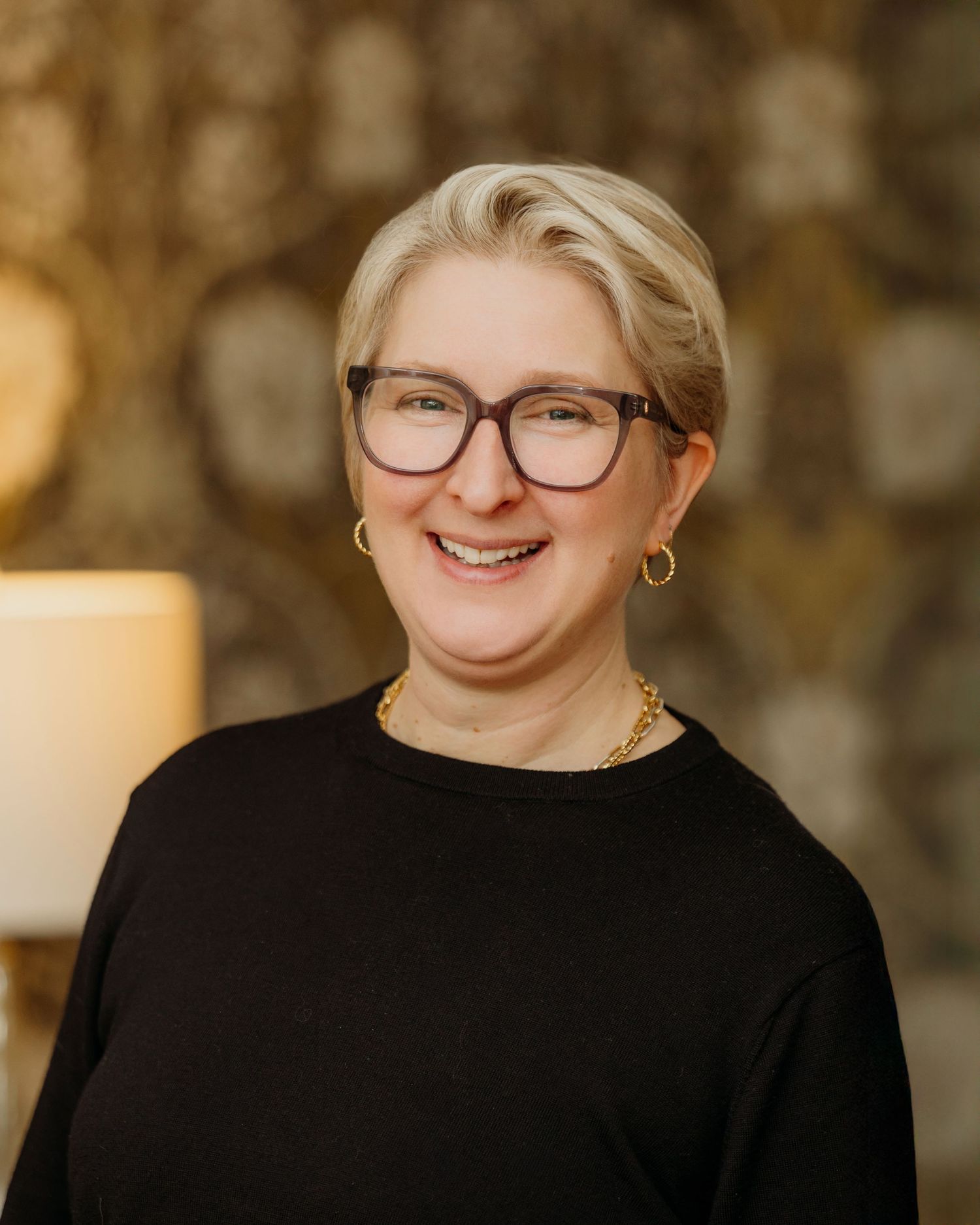 Portrait of person in black top wearing glasses against warm toned background.