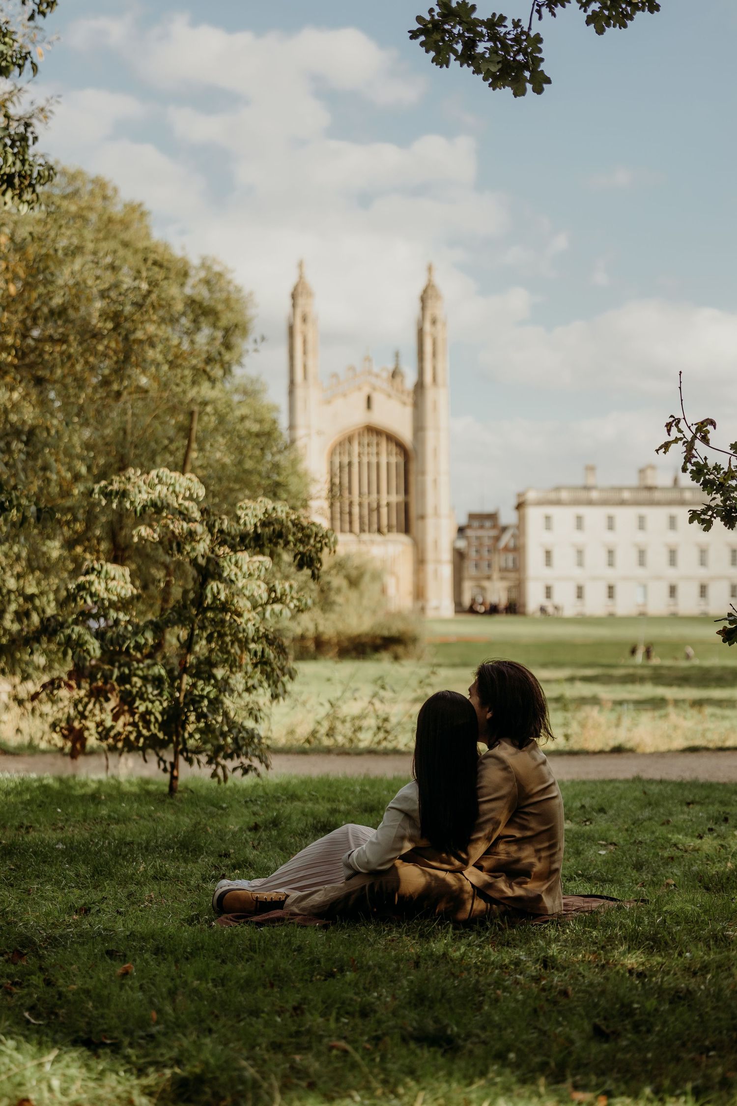 A couple relaxes on grass with Cambridge University's iconic chapel towers in the background.