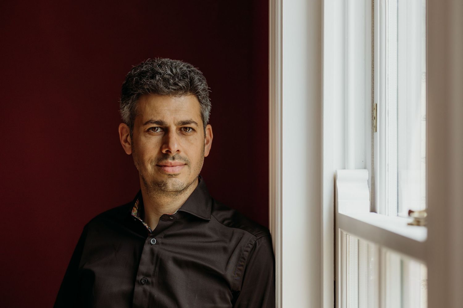 Professional headshot of someone in a black shirt against a red wall and window.