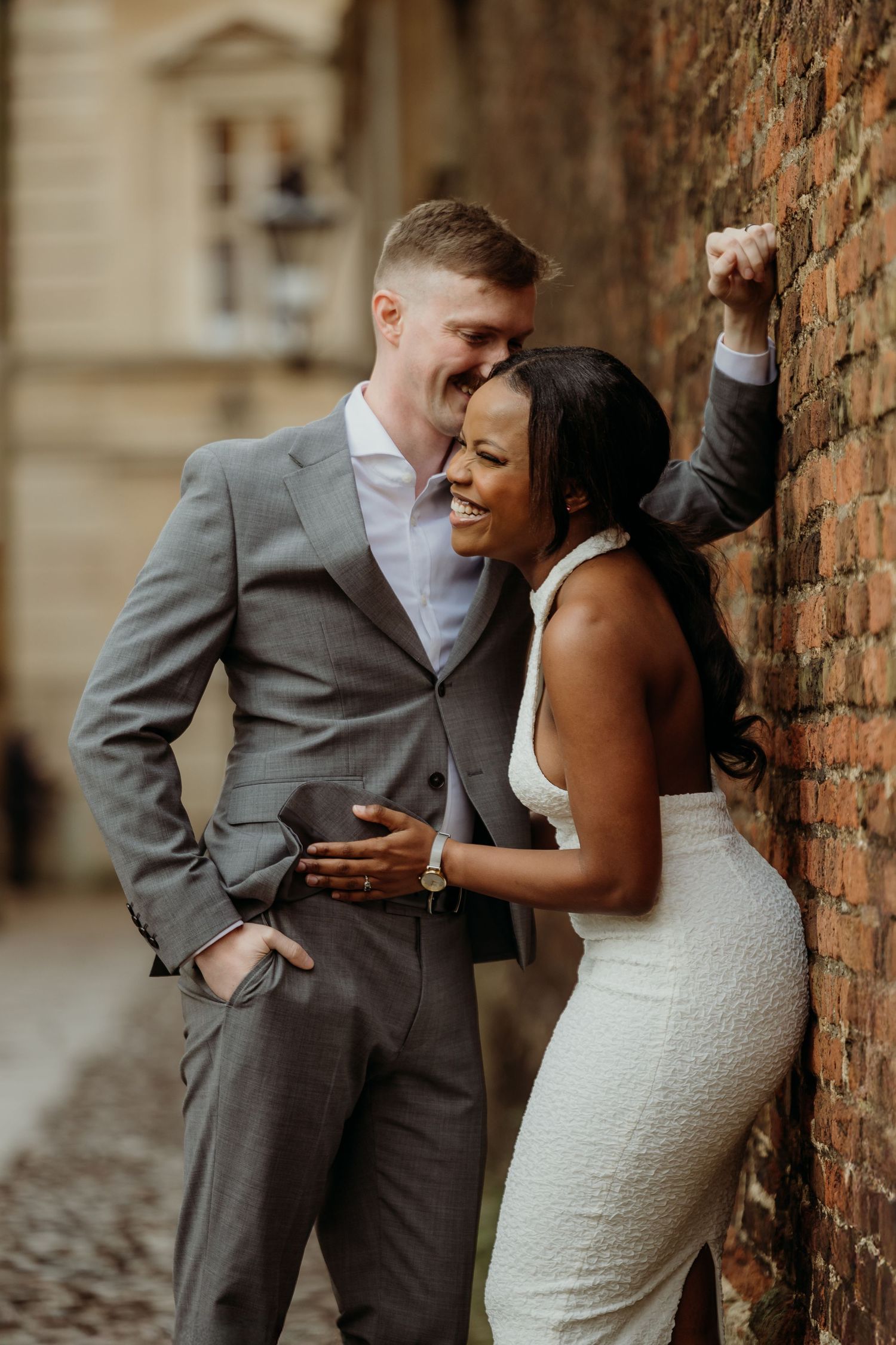 A couple in wedding attire shares playful moments against a brick wall.