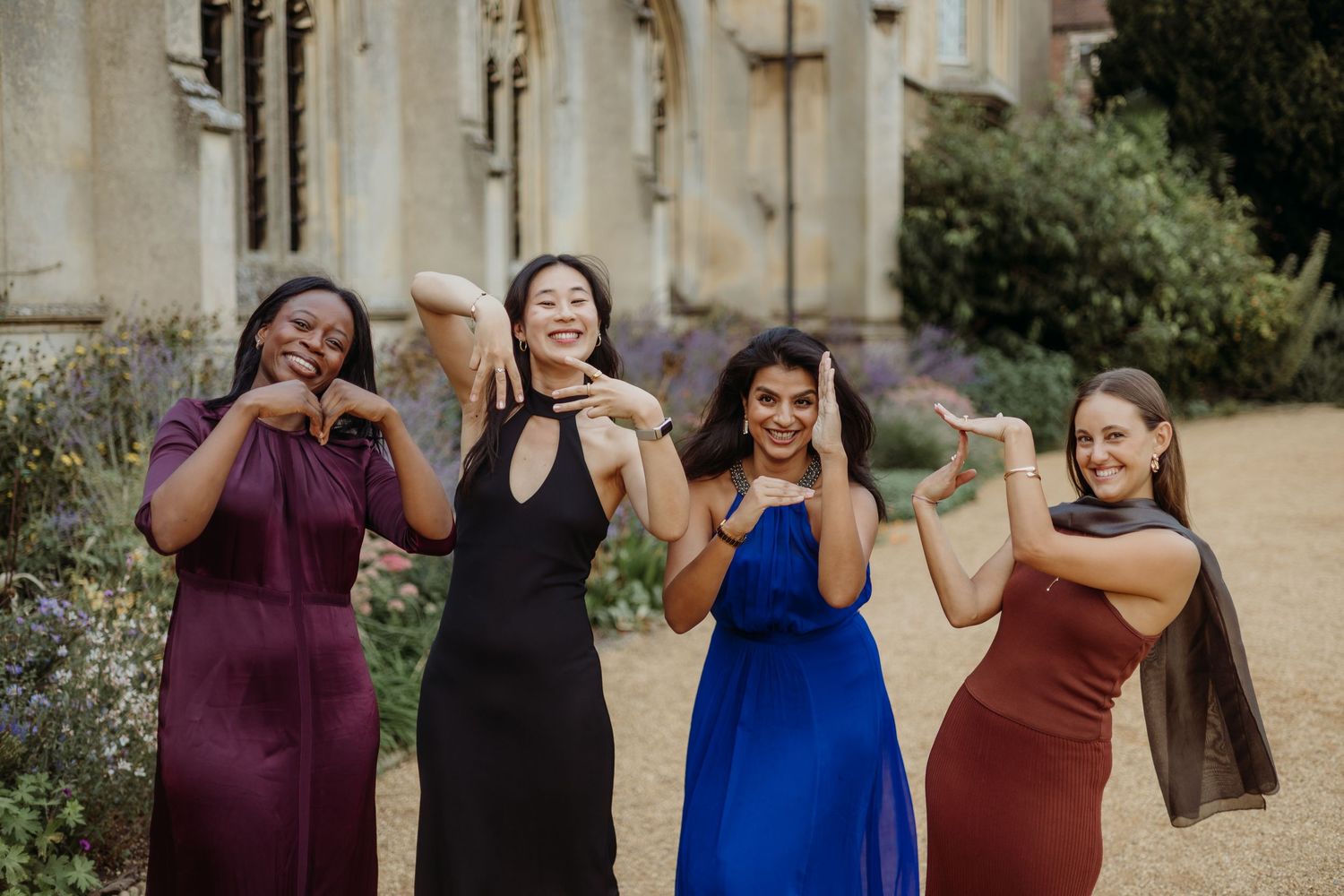 Four women in elegant dresses pose playfully outside a historic building.