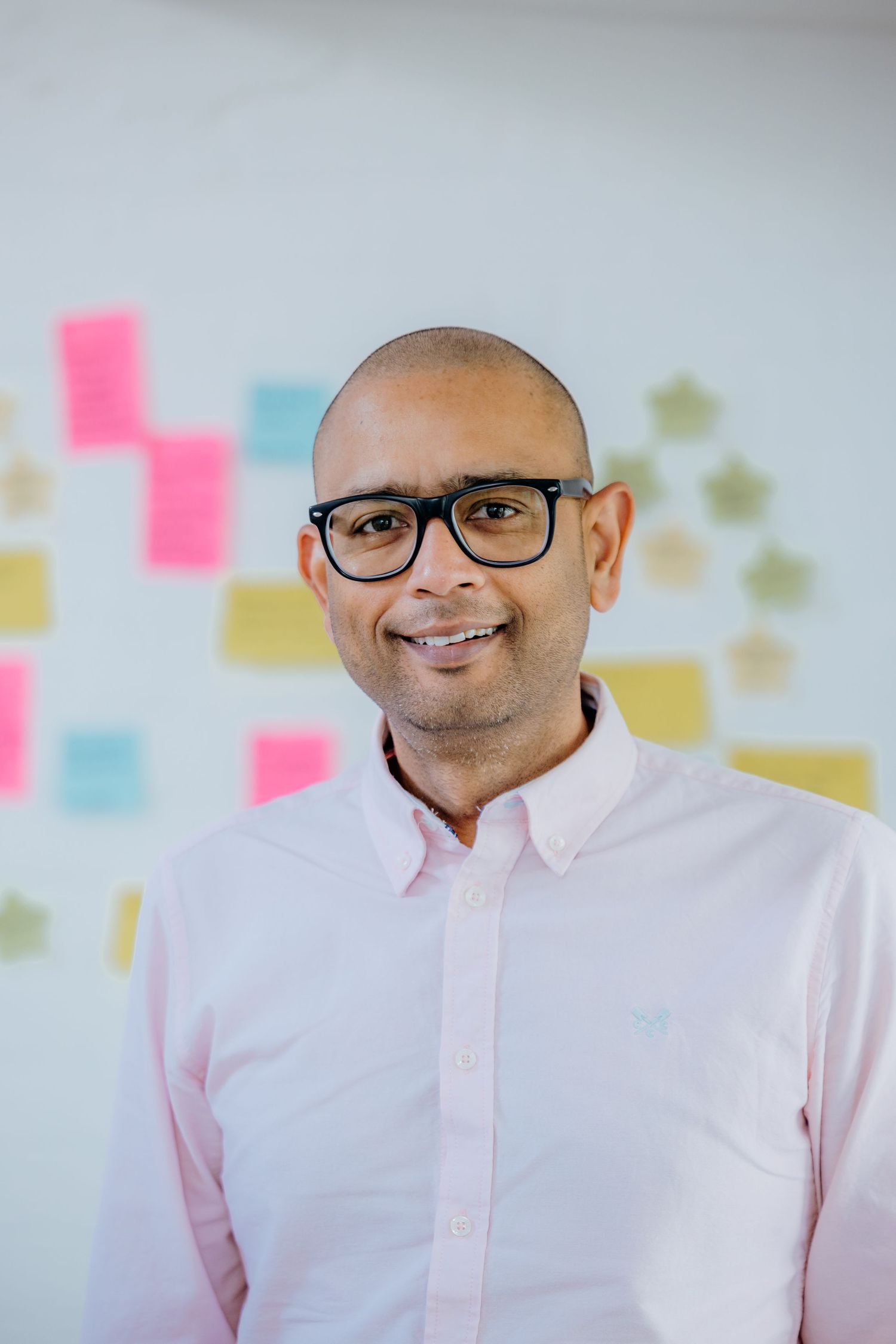 Professional headshot of person in pink shirt and glasses against light background with sticky notes.