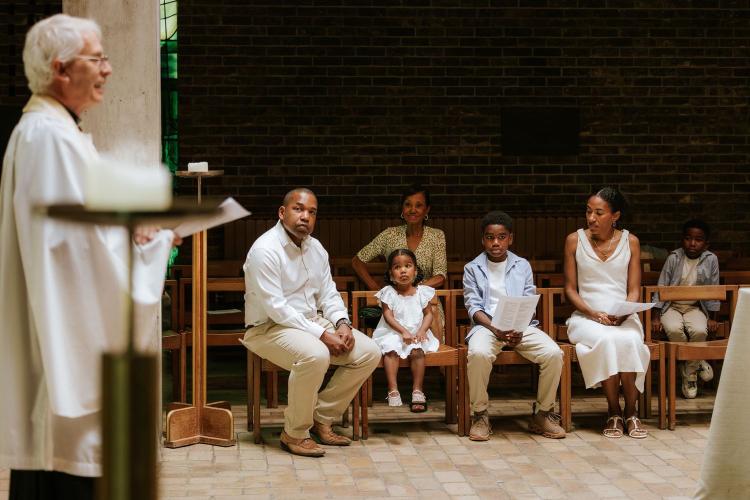 People seated in white clothing along a dark wall during a ceremony.