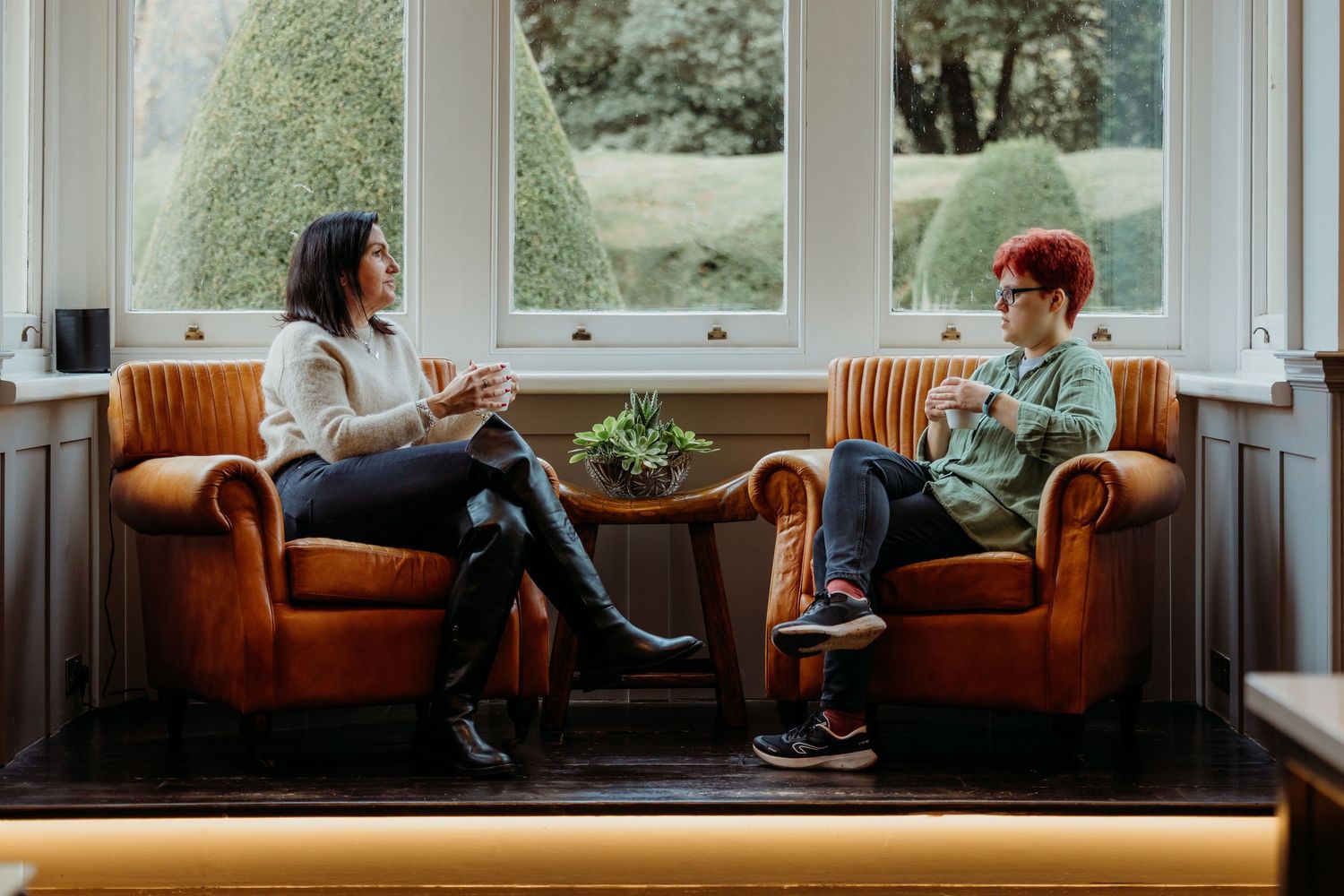 Two people having a casual conversation while sitting on orange armchairs by a window.