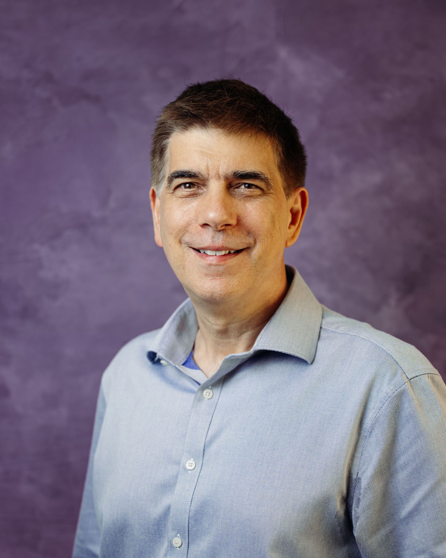 Professional headshot of person in light blue collared shirt against purple background.