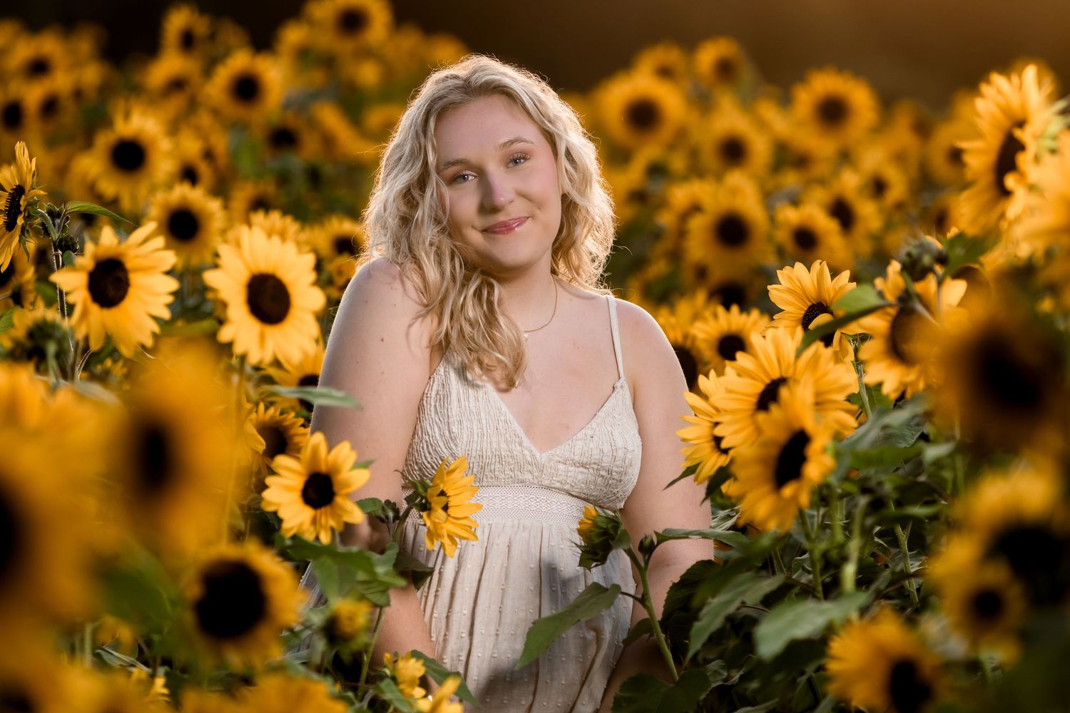 Smiling blonde woman in white dress surrounded by vibrant yellow sunflowers in a sunlit field.