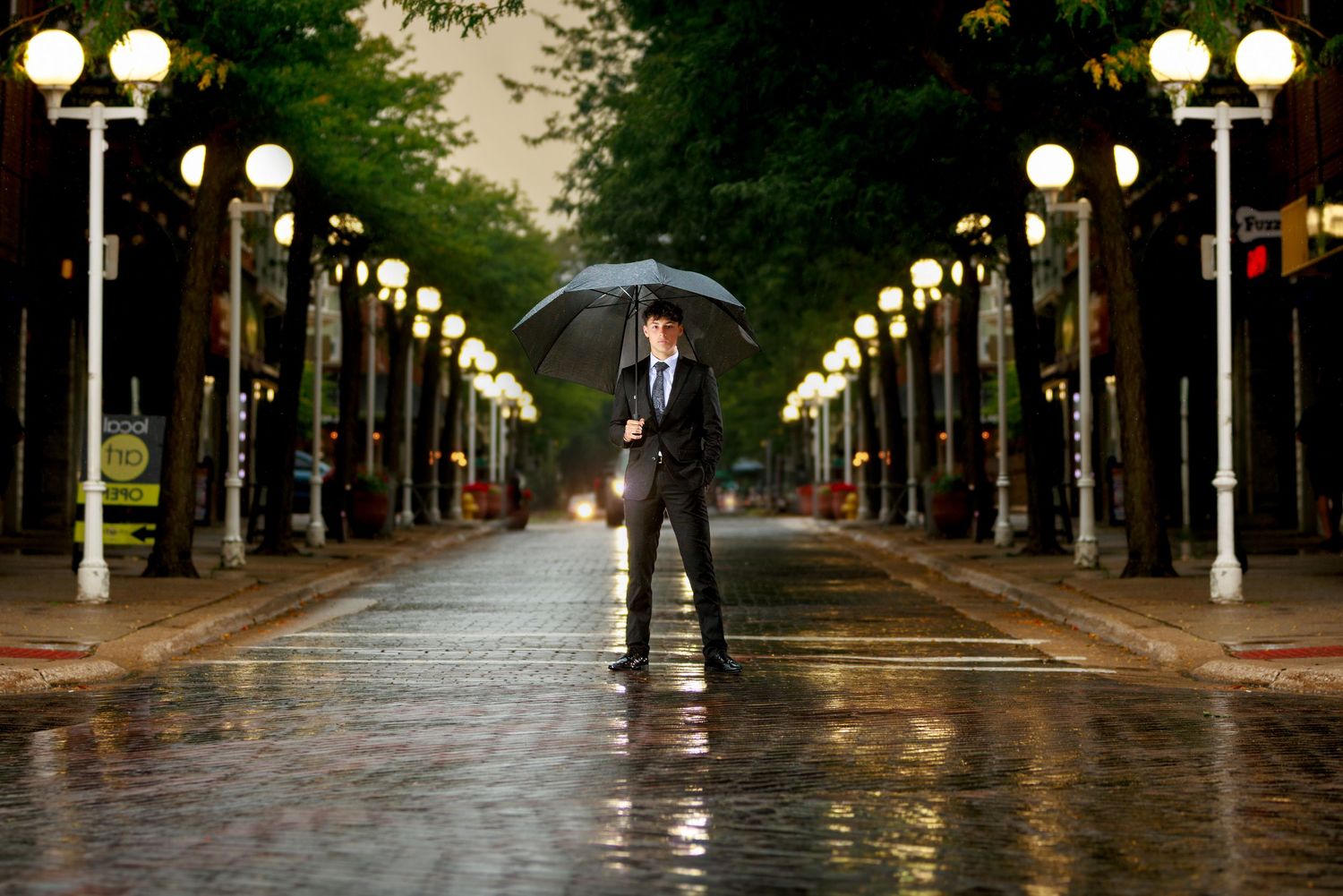 Man in suit holding umbrella standing on rain-soaked cobblestone street lined with glowing lamp posts.