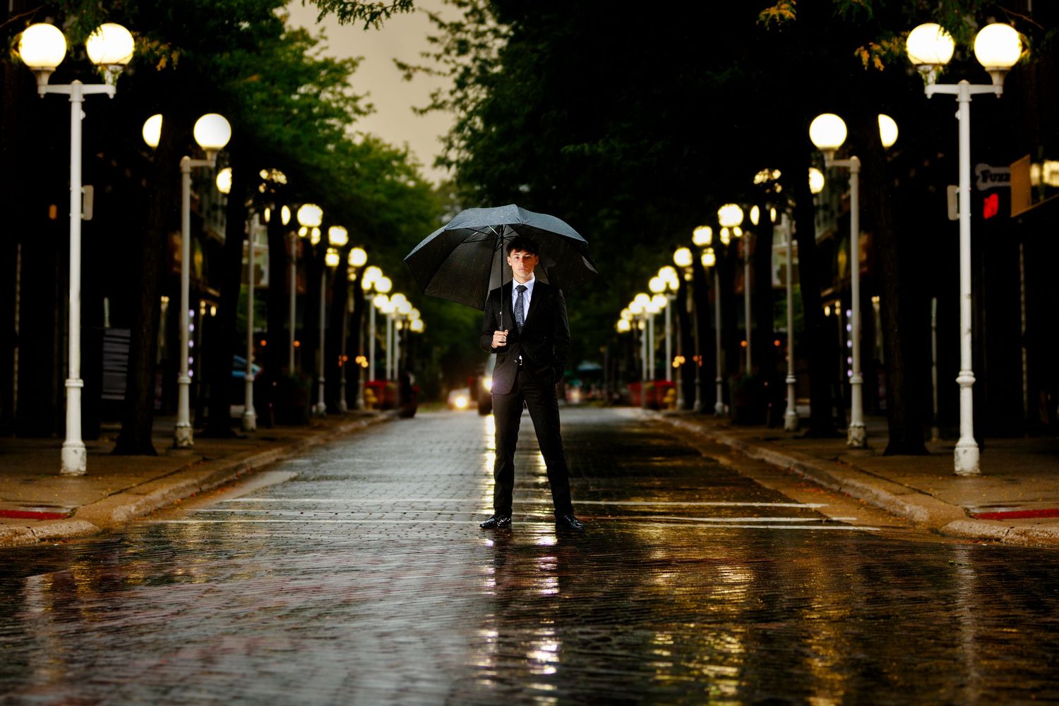 A man in a suit holds an umbrella on a rain-soaked street lined with glowing lamp posts at night.