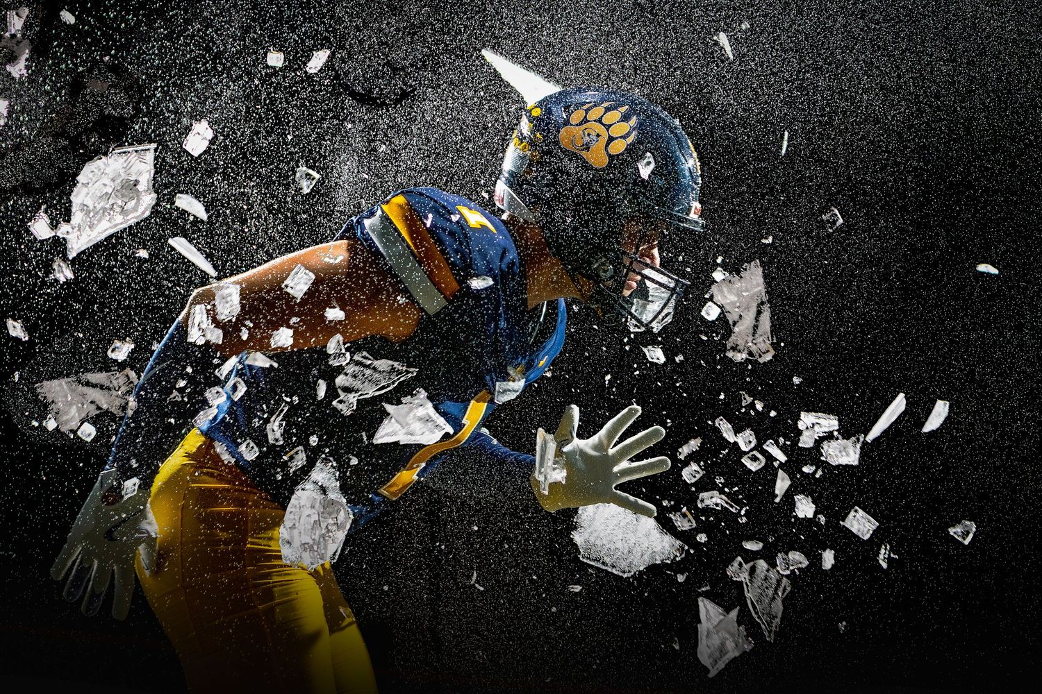 American football player in blue and gold uniform bursting through shattering glass on dark background.