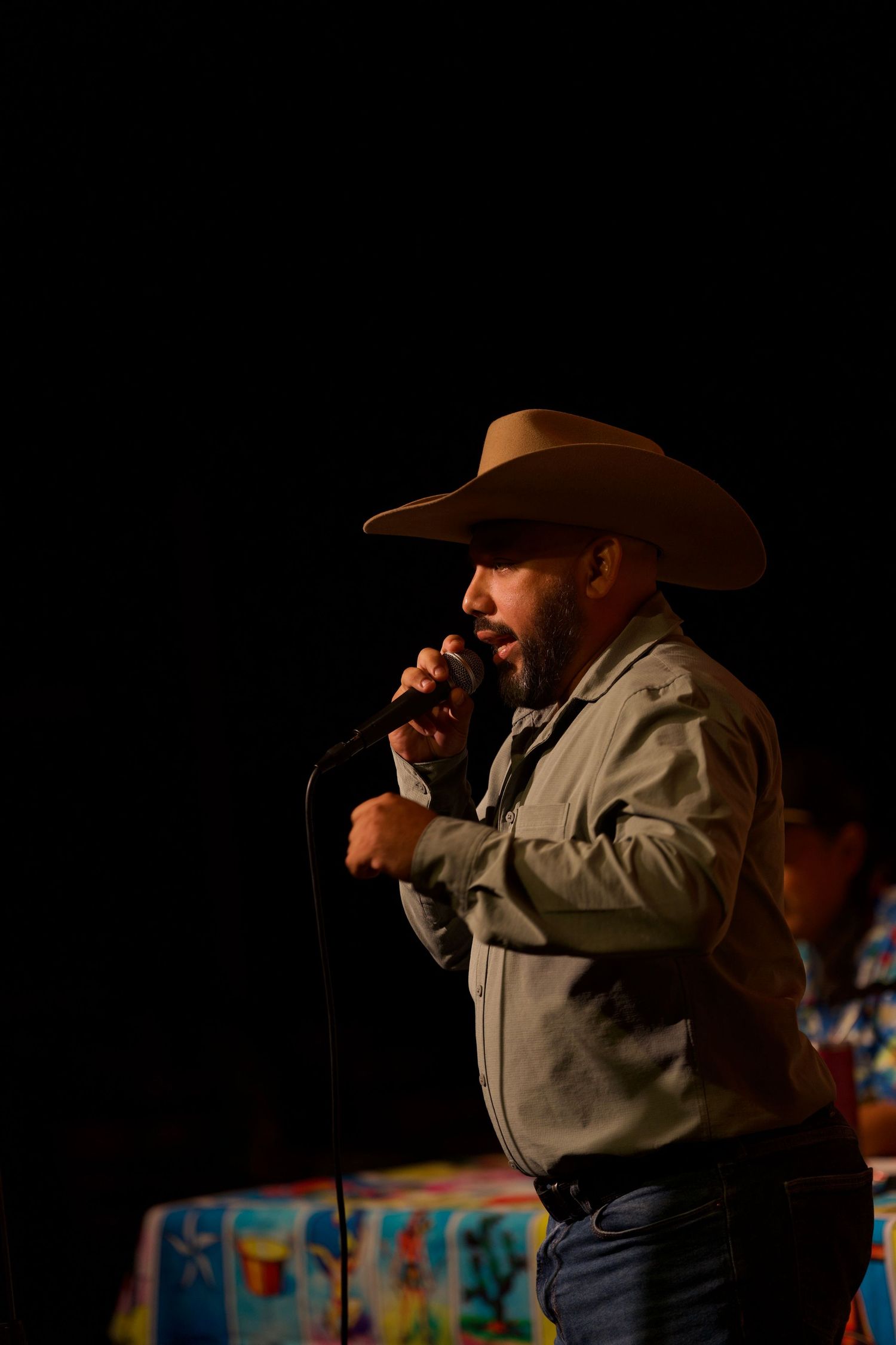 Performer in cowboy hat and tan jacket delivers standup routine on dark stage.