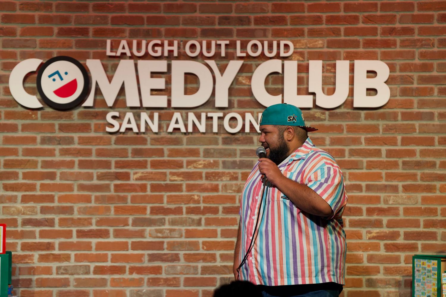Comedy club stage with brick wall and 'Laugh Out Loud' signage visible in background.