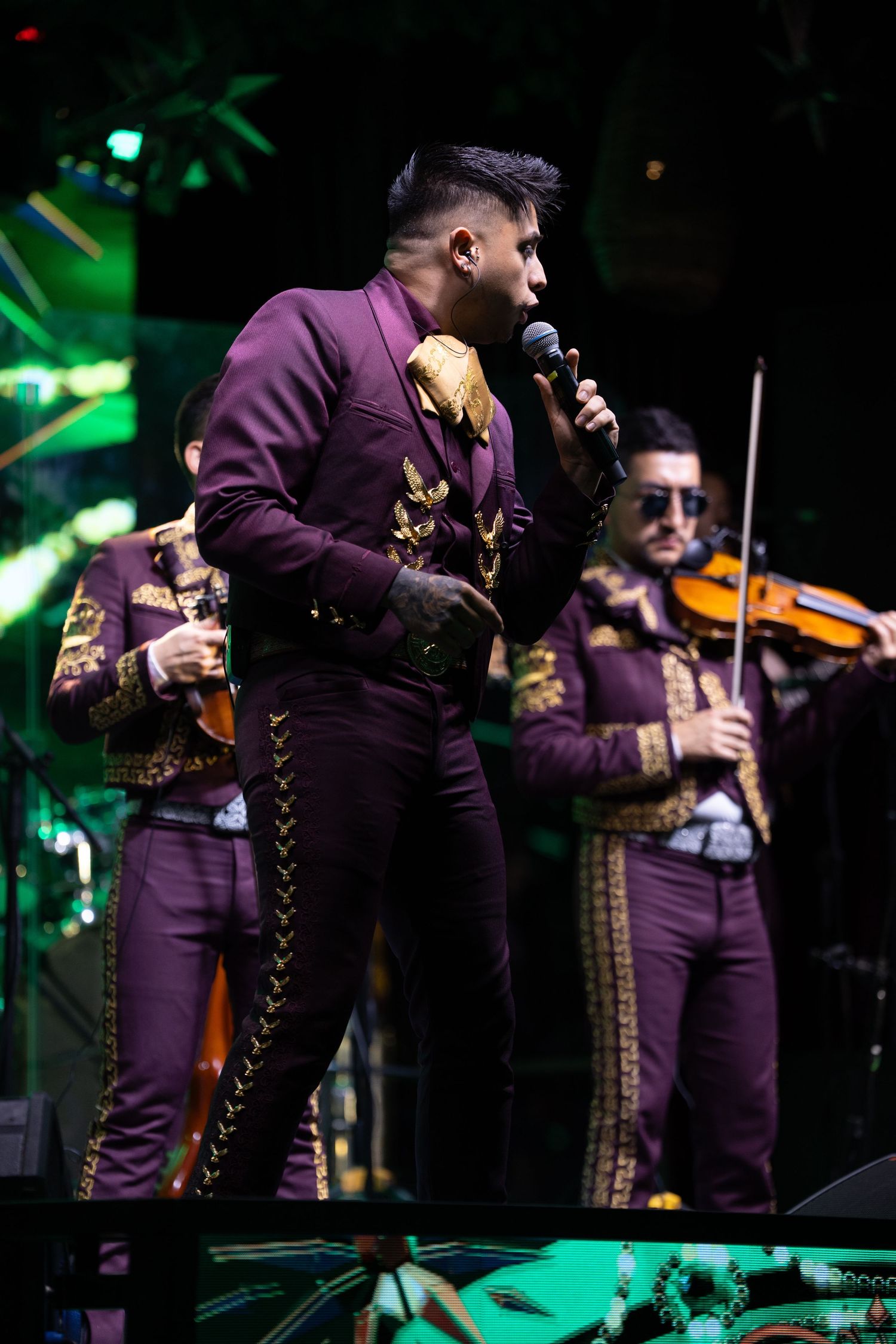 Mariachi performer in purple suit singing on stage with violin players in background.