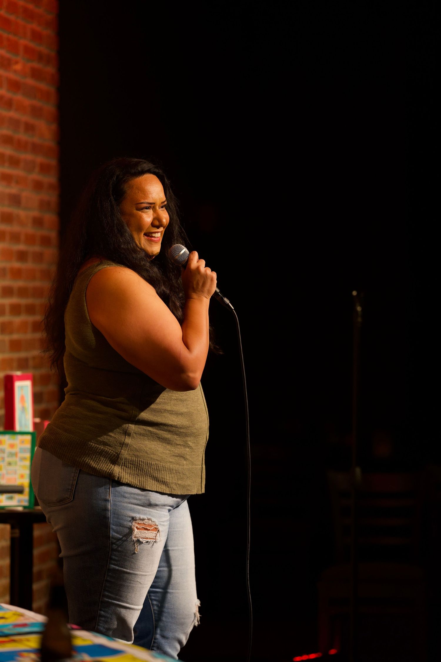 Performer in olive tank top and jeans delivering stand-up comedy at a brick-walled venue.