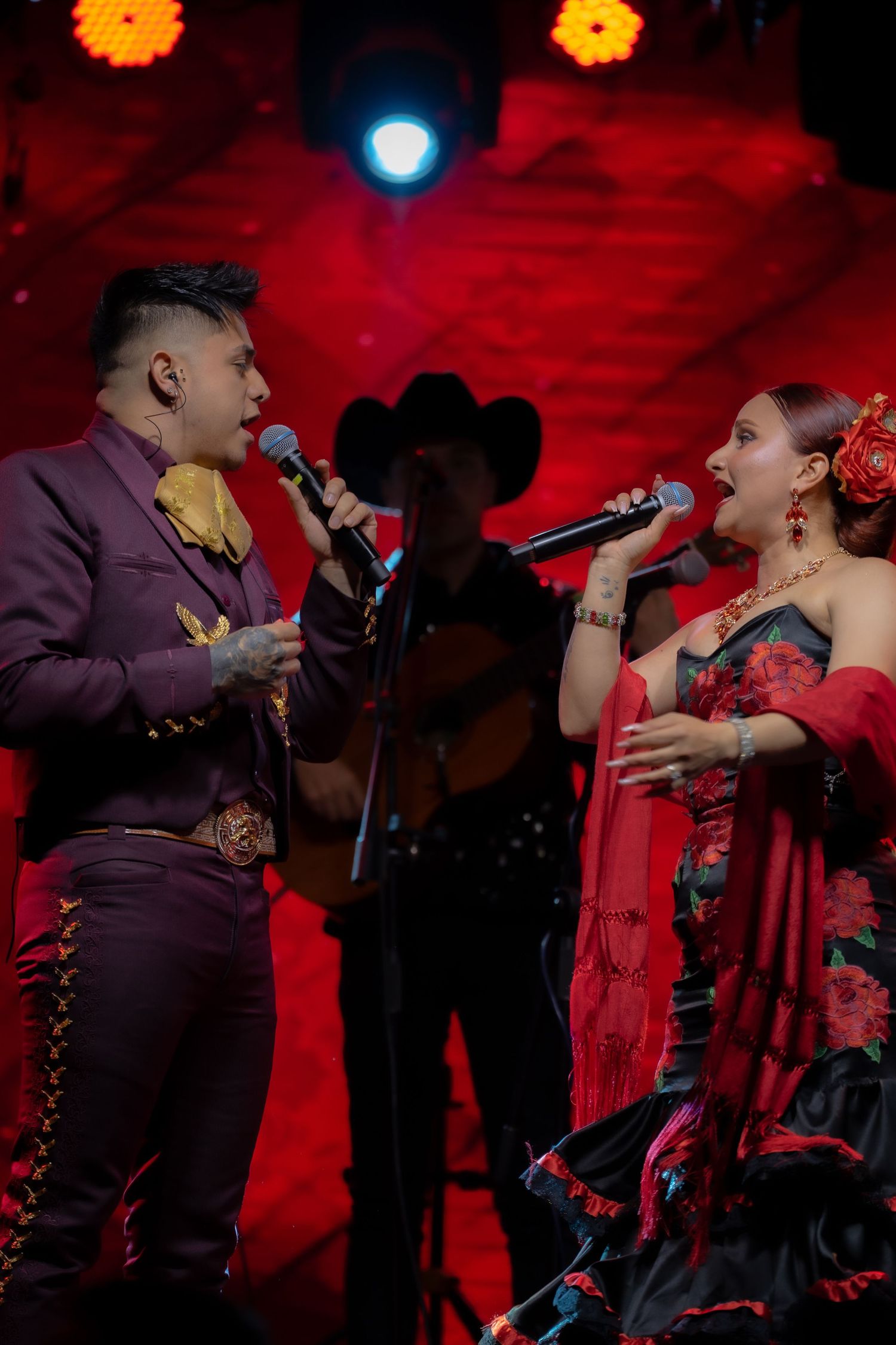 Performers singing a duet on stage with vibrant red lighting and mariachi band in background.