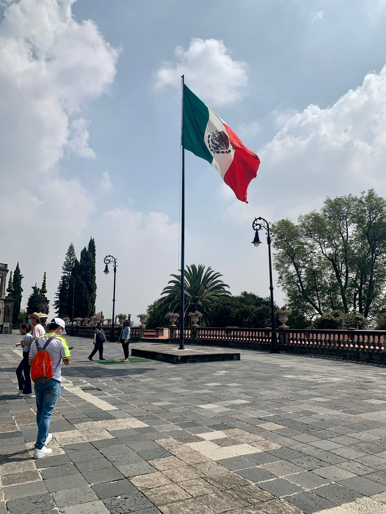 Stone plaza with Mexican flag waving against blue sky and palm trees.