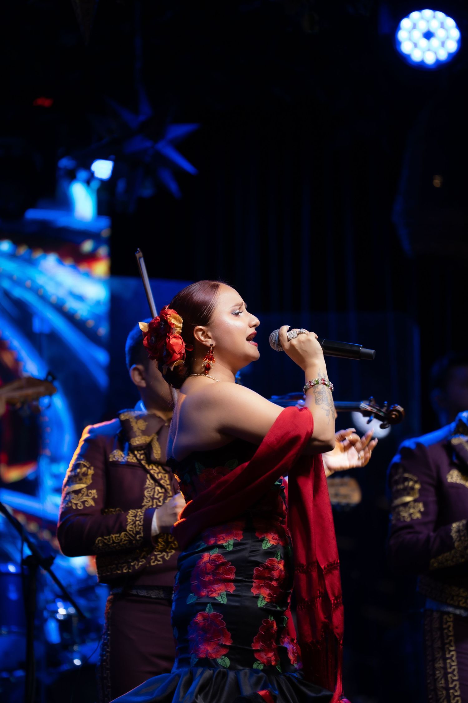 Performer in traditional Mexican dress singing passionately on stage with dramatic blue lighting.