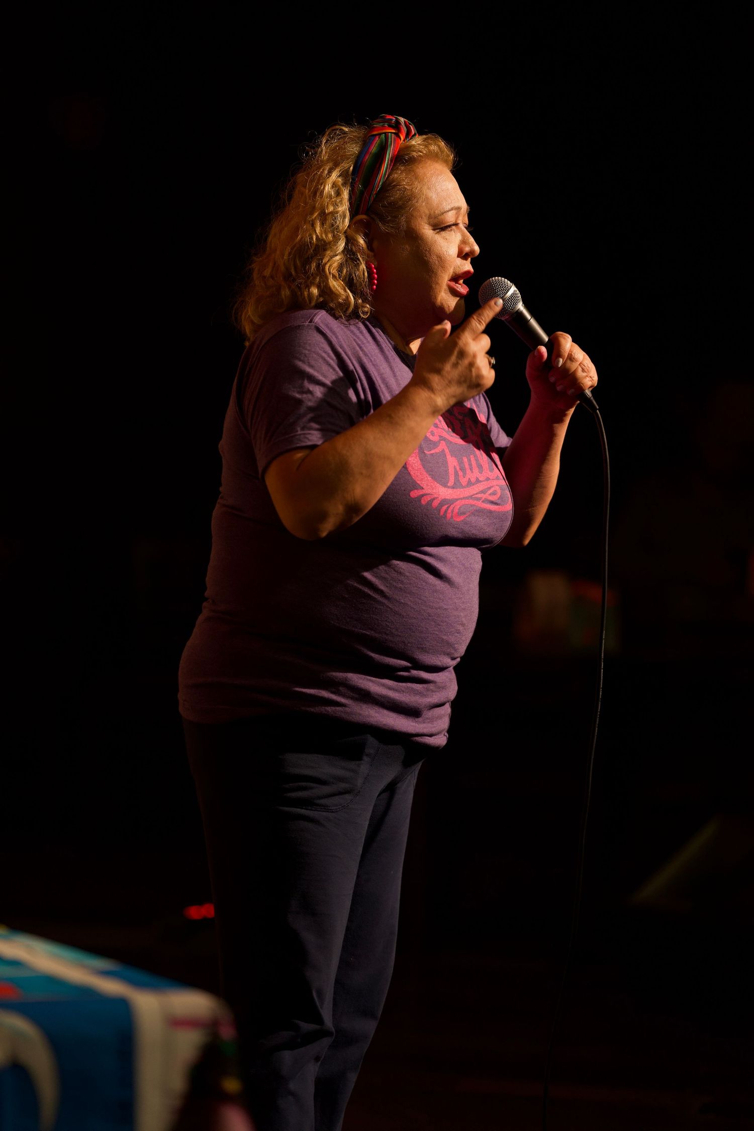 Performer in purple shirt delivers standup routine on dimly lit comedy stage.