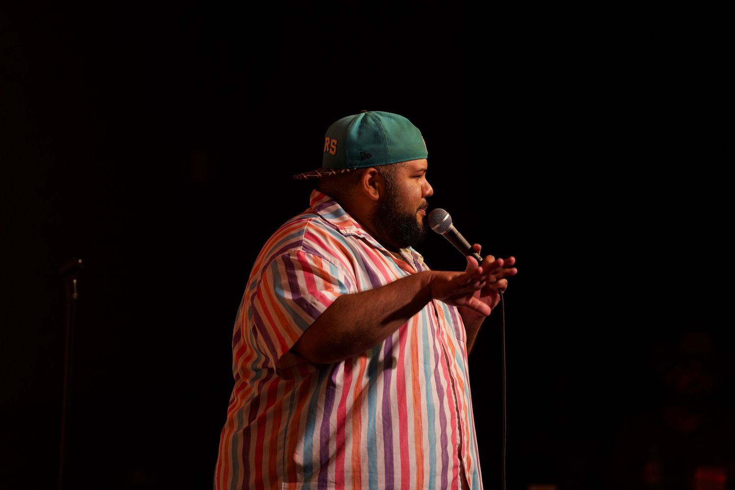 Comedian in striped shirt performs on stage at San Antonio comedy club against brick wall backdrop.