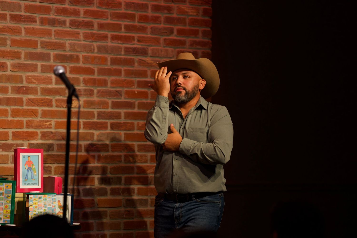 Comedian in western attire performs against brick wall at comedy club venue.