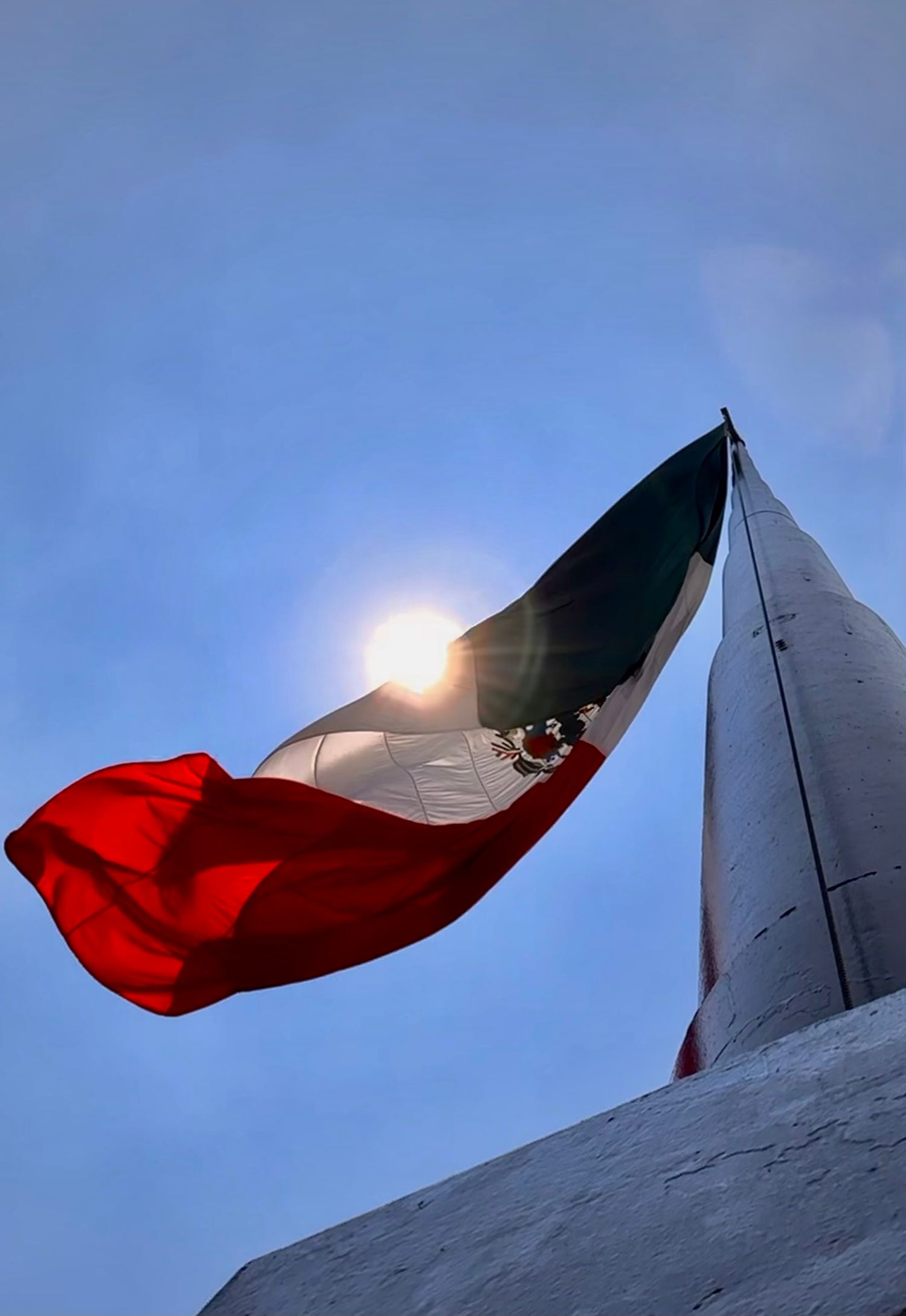 Mexican flag waving against blue sky with sun creating dramatic backlighting.