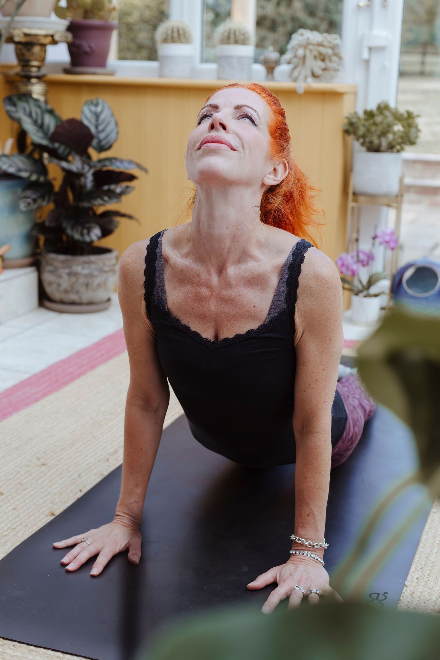 A woman with red hair practices yoga indoors, lifting her chest and looking upward in a strong, focused backbend on her mat.