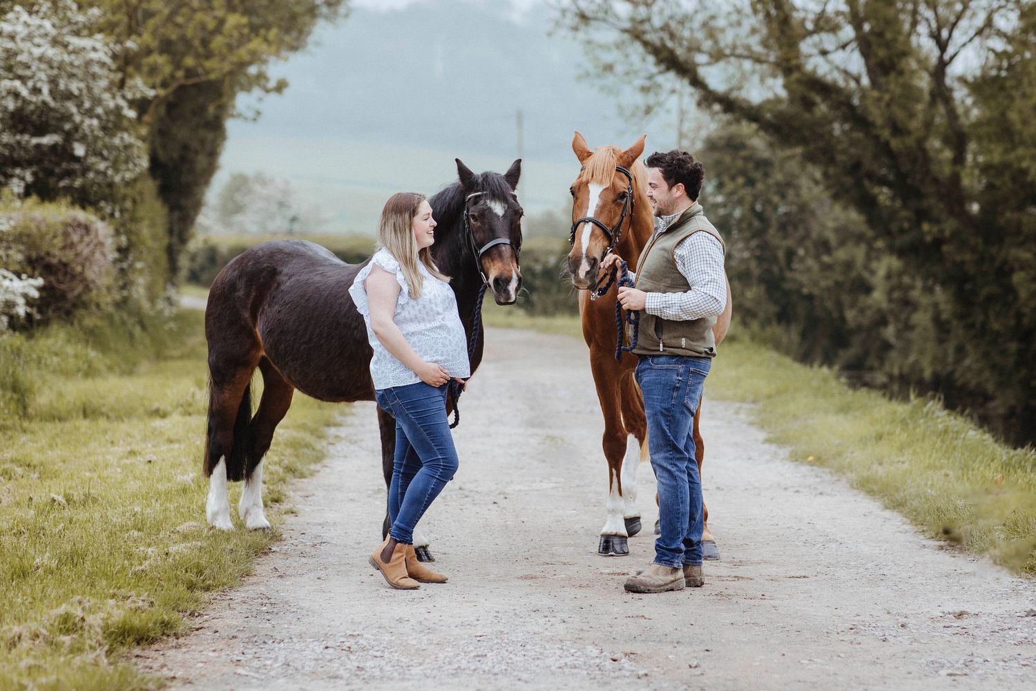 A couple stands on a country path with two horses, sharing a calm moment surrounded by greenery.